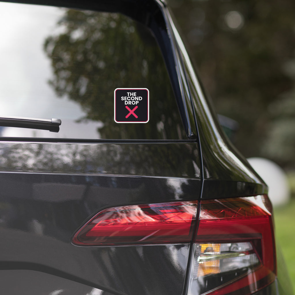 Car with a colorful sticker on the rear window, blurred greenery in the background The Second Drop with a red X