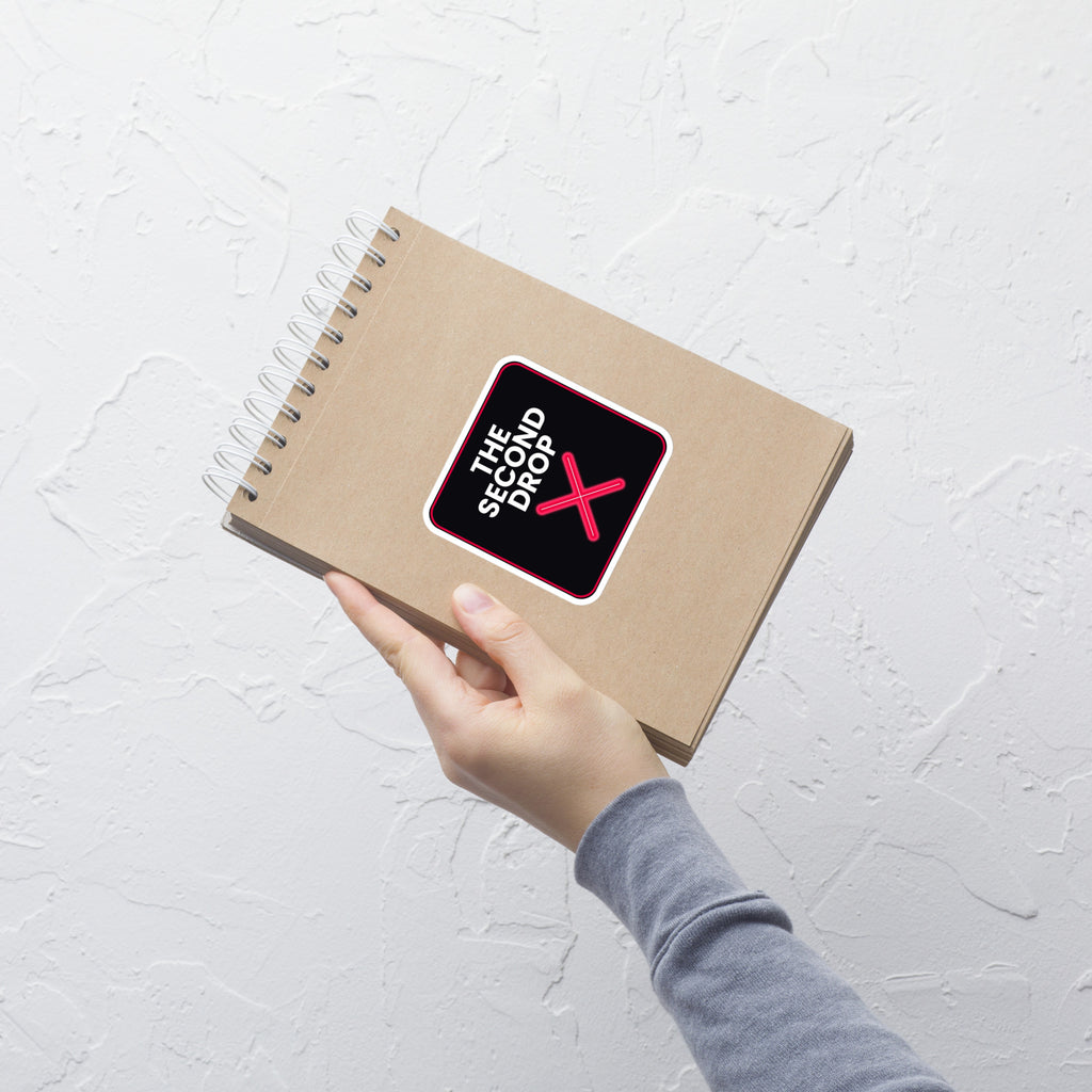 A hand holding a brown kraft paper spiral notebook against a textured white wall. The notebook cover features a black square sticker with a pink border and the text 'THE SECOND DROP X' in white and red