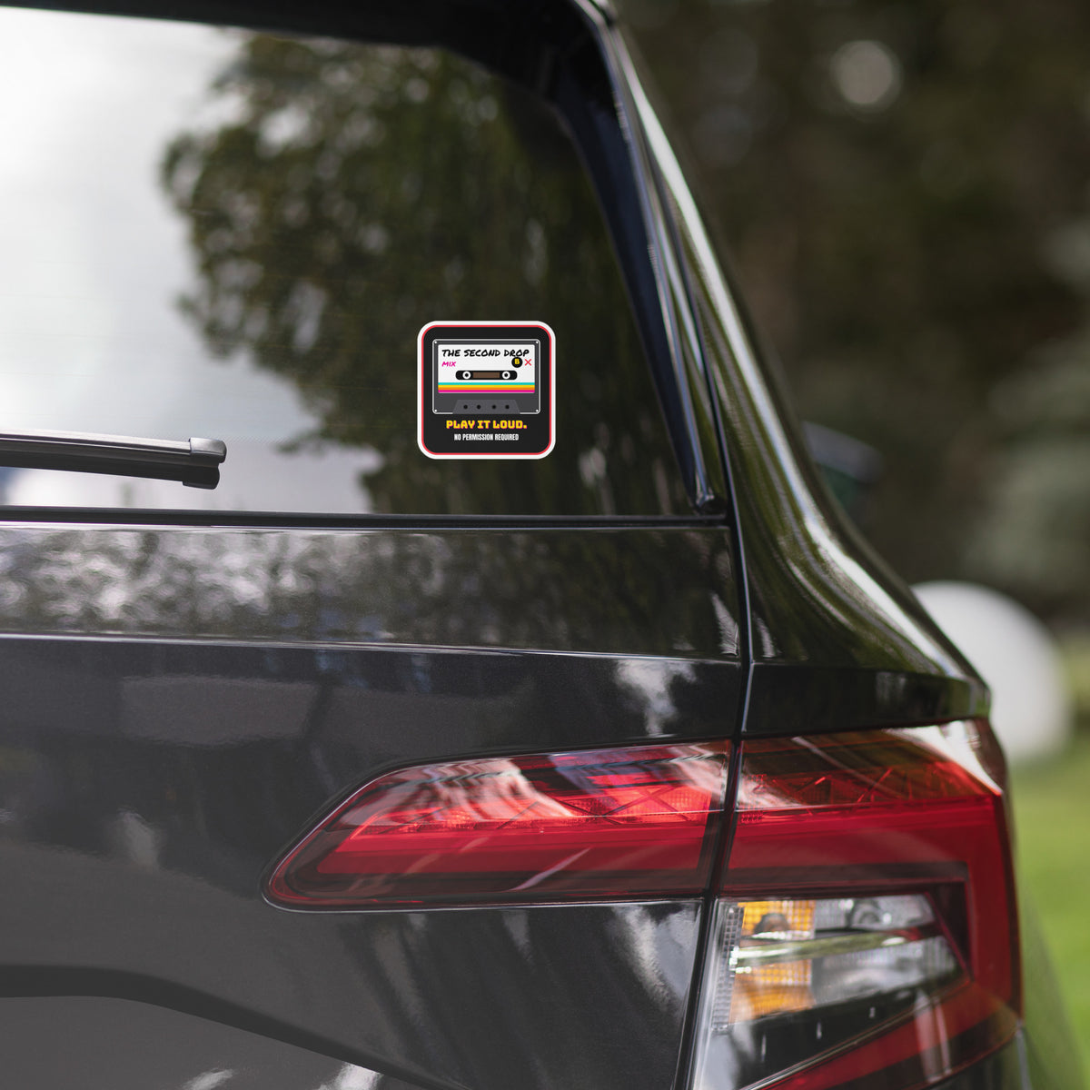 Car with a small square decal on the back window. A square sticker or decal with rounded corners featuring a retro cassette tape graphic. The design has a black background with a red border, a white cassette face, and colorful rainbow stripes. The Second Drop. Play It Loud. No Permission Required. 