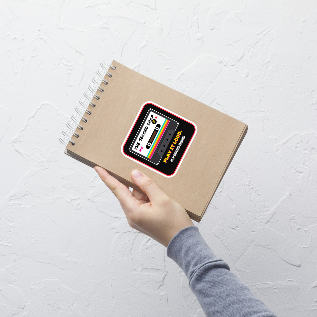 A hand holding a spiral-bound notebook with a retro cassette tape graphic on the cover, set against a textured white wall. A square sticker or decal with rounded corners featuring a retro cassette tape graphic. The design has a black background with a red border, a white cassette face, and colorful rainbow stripes. The Second Drop. Play It Loud. No Permission Required. 