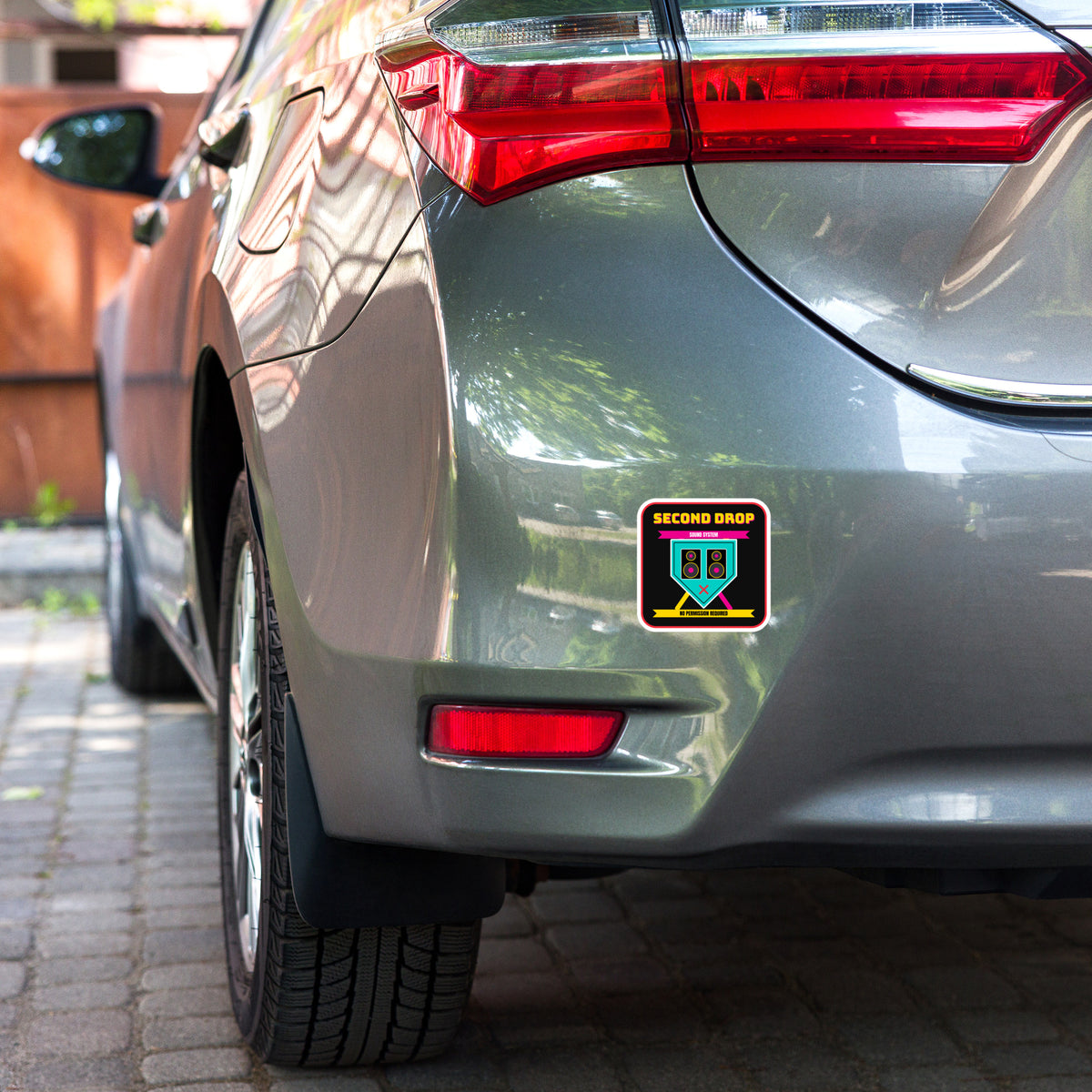 Car with a sticker with A square sticker or decal with rounded corners featuring a black background and a colorful graphic. The design shows the text 'SECOND DROP SOUND SYSTEM' in yellow and pink, a teal speaker-shaped emblem with speakers and a pink X, and yellow text reading 'NO PERMISSION REQUIRED' on a white border.