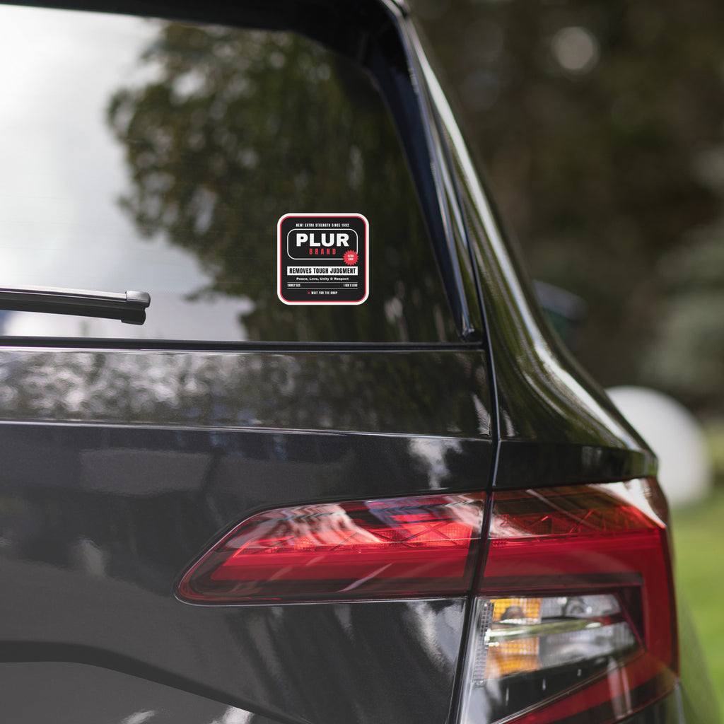 A small rectangular pink and white PLUR car window sticker applied to the rear window of a black SUV, with a blurred outdoor background of trees and grass.