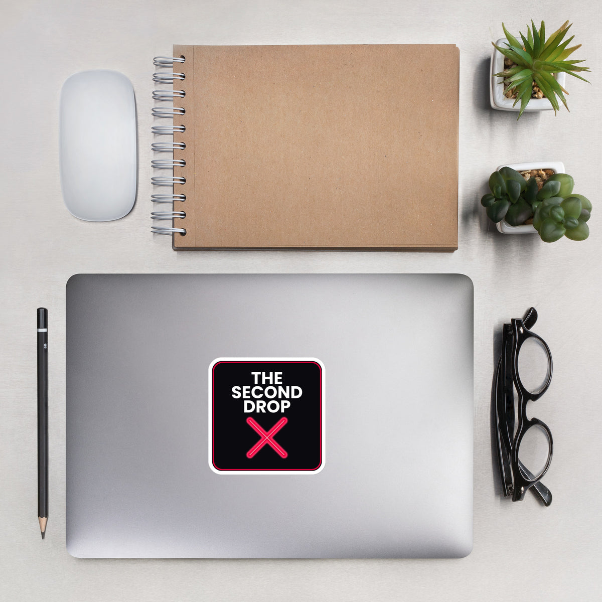 The Second Drop with red X on a Laptop with a decorative sticker on a desk with a notebook, pens, and a plant.