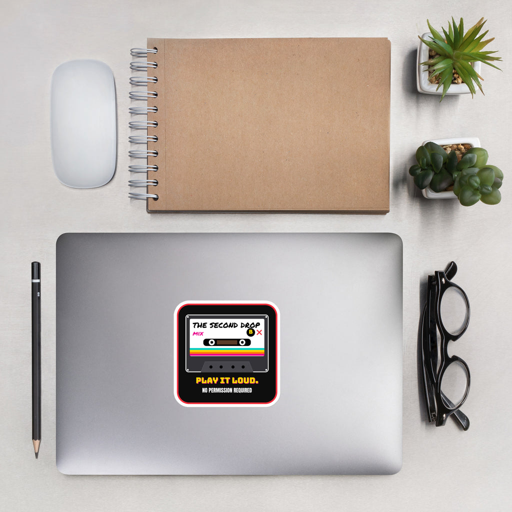 A square sticker with a retro cassette tape design is applied to the center of a silver laptop. A square sticker or decal with rounded corners featuring a retro cassette tape graphic. The design has a black background with a red border, a white cassette face, and colorful rainbow stripes. The Second Drop. Play It Loud. No Permission Required. 