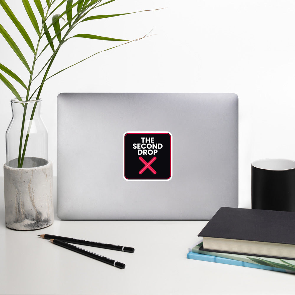 The Second Drop with red X on a Laptop with a decorative sticker on a desk with a notebook, pens, and a plant.