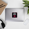 The Second Drop with red X on a Laptop with a decorative sticker on a desk with a notebook, pens, headphones, and a plant.