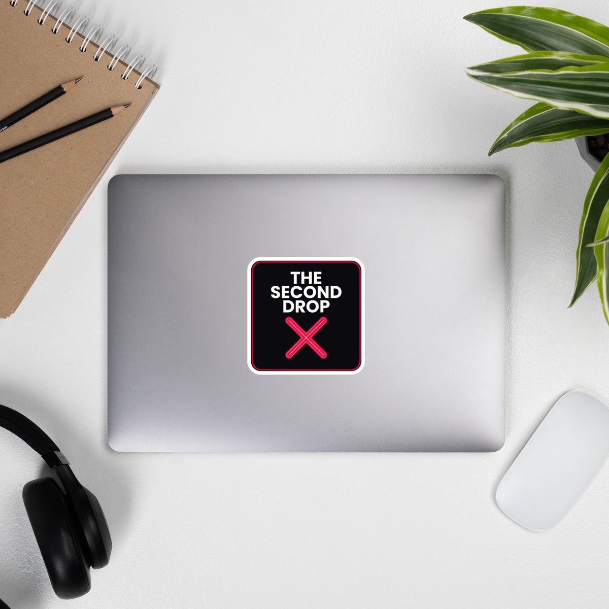 The Second Drop with red X on a Laptop with a decorative sticker on a desk with a notebook, pens, headphones, and a plant.