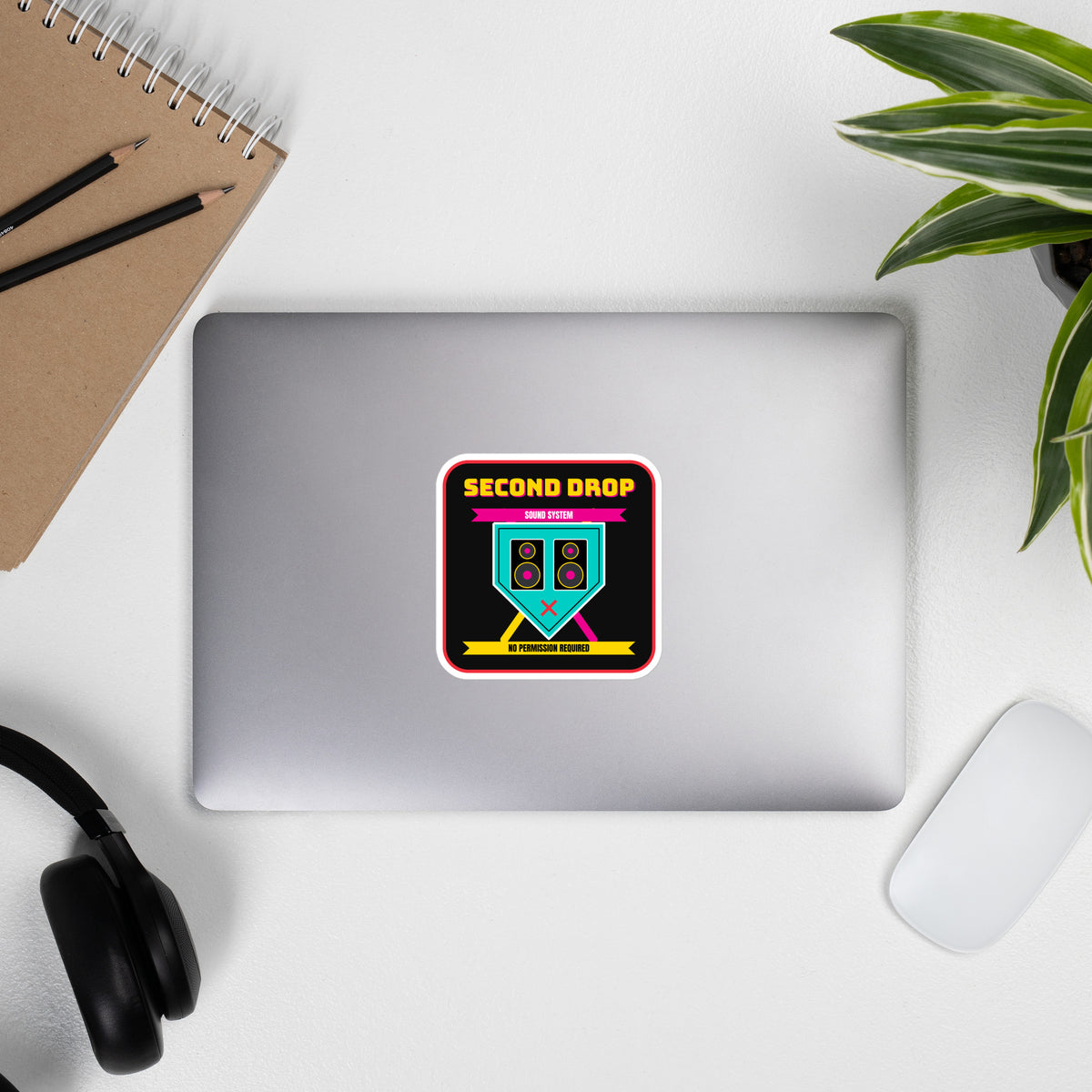 A square sticker or decal with rounded corners featuring a black background and a colorful graphic. The design shows the text 'SECOND DROP SOUND SYSTEM' in yellow and pink, a teal speaker-shaped emblem with speakers and a pink X, and yellow text reading 'NO PERMISSION REQUIRED' on a white border. The laptop is photographed from above on a white desk with a notebook, headphones, mouse, and plant around it.