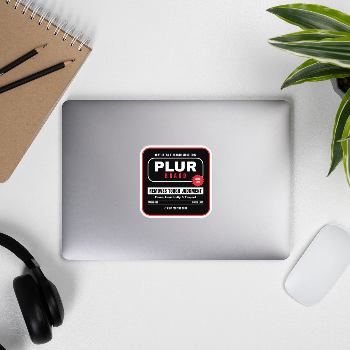 A silver laptop on a white desk with a rectangular sticker that reads 'PLUR  in the center. The scene is styled as a flat lay with a brown notebook, pencils, black headphones, a white mouse, and a green plant.