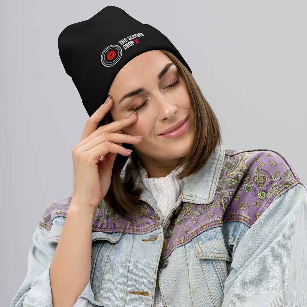 Woman wearing a black beanie with text THE SECOND DROP and a patterned denim jacket on a gray background
