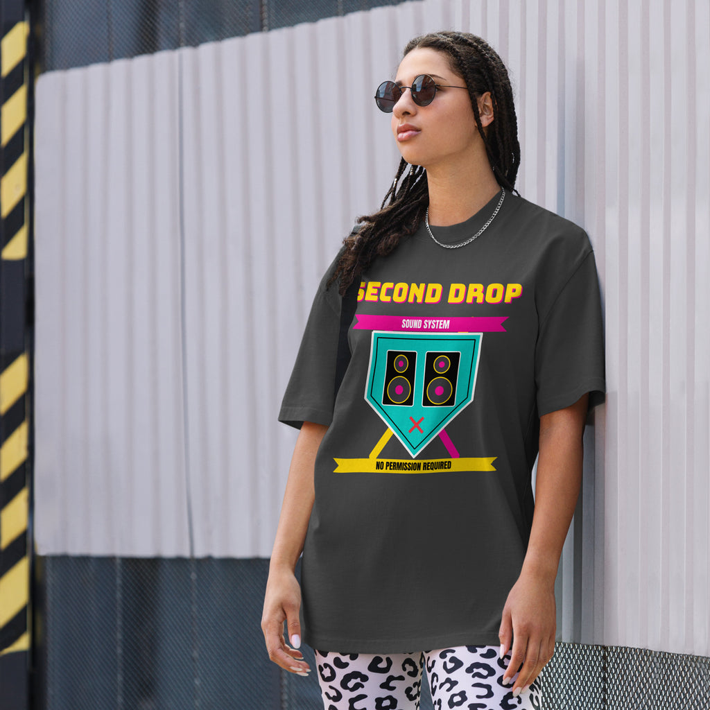 A woman wearing a dark charcoal gray graphic t-shirt with a retro-style 'SECOND DROP DRUM SYSTEM' design in yellow, pink, and teal, standing against a corrugated metal wall.